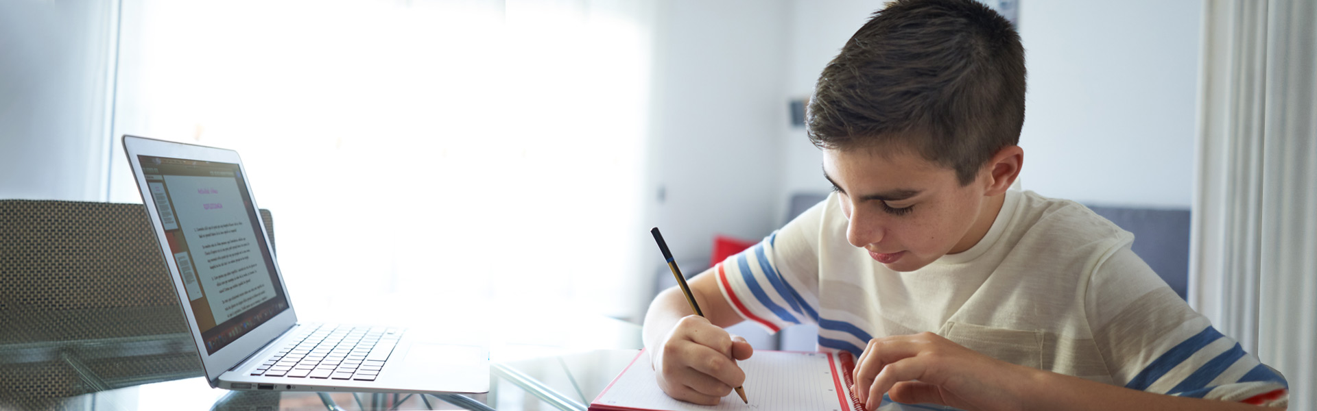 Young boy sitting at table with notepad and laptop doing homework