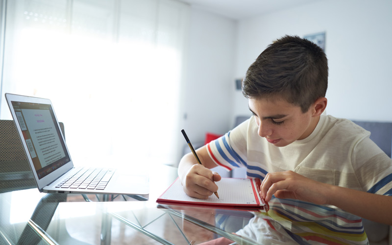 Young boy sitting at table with notepad and laptop doing homework