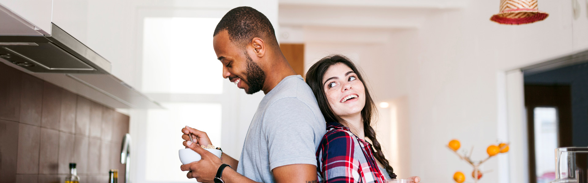 Man eating bowl of cereal with woman leaning against his back smiling at him