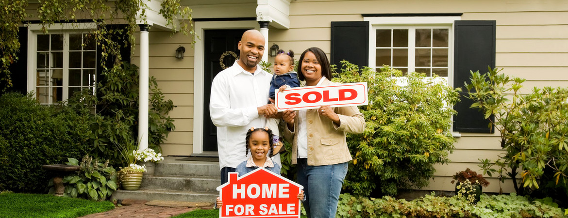 Young family of four holding sold and home for sale signs in front of their new home